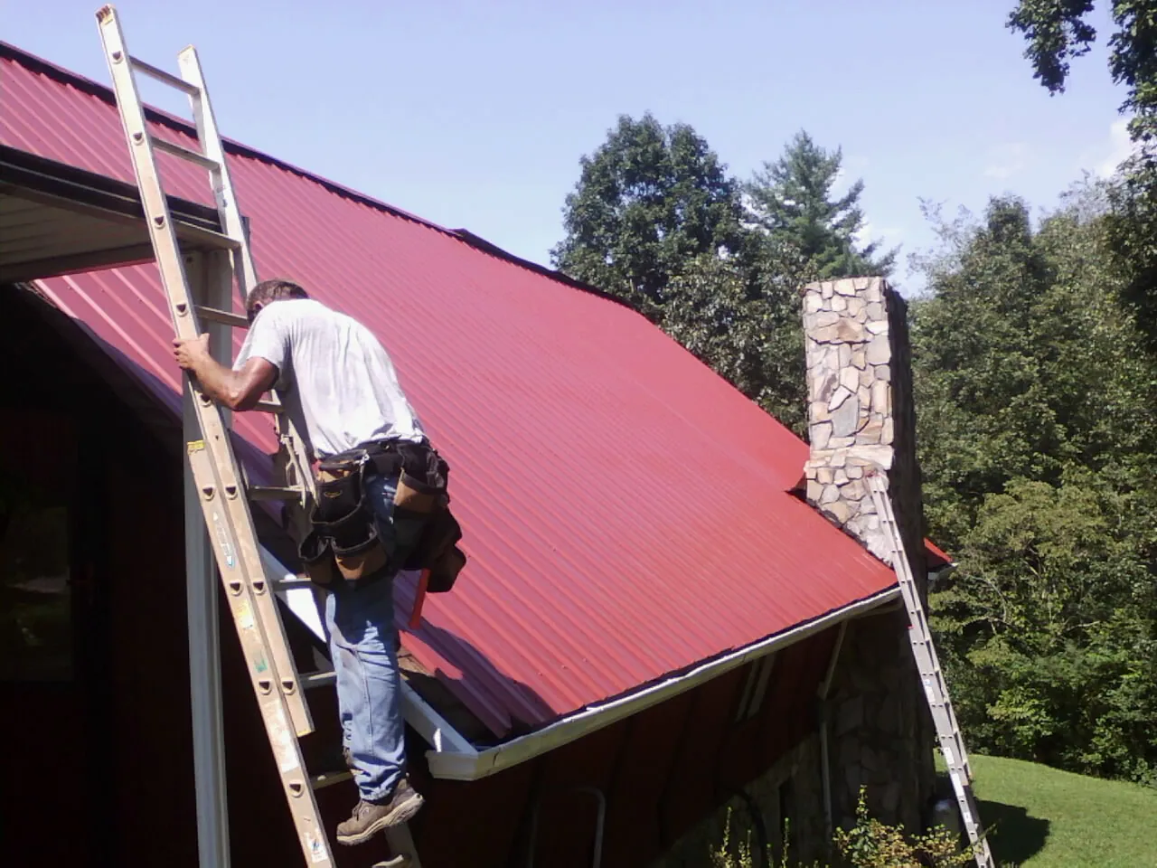 Roofer on a ladder beside a red metal roof and stone chimney — Soffit & Fascia Repair by Roofing Brothers in Louisburg